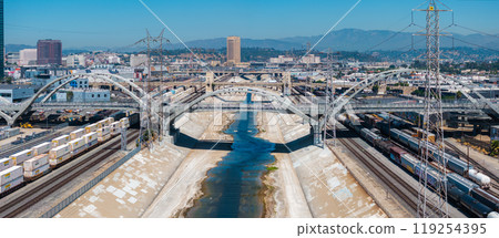 The iconic Sixth Street Viaduct spans the Los Angeles River, surrounded by industrial areas and train tracks. The cityscape and mountains are visible under a clear sky. The iconic Sixth Street Viaduct spans the Los Angeles River, surrounded by industrial areas and train tracks. The cityscape and mountains are visible under a clear sky. 119254395