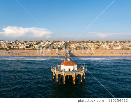 The iconic Manhattan Beach Pier with a red roofed building extends into the Pacific Ocean, surrounded by sandy beaches and a densely packed residential area. The iconic Manhattan Beach Pier with a red roofed building extends into the Pacific Ocean, surrounded by sandy beaches and a densely packed residential area. 119254412