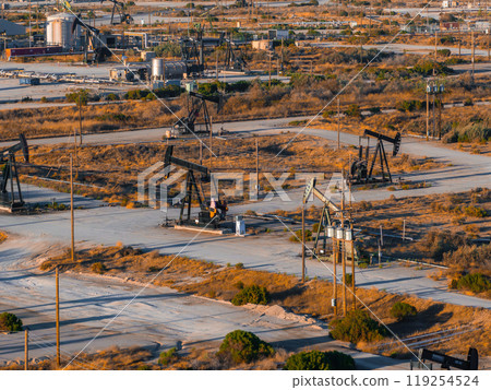Numerous oil rigs operate in a desert landscape in California, with dusty roads and sparse vegetation. The setting sun casts long shadows over the scene. Numerous oil rigs operate in a desert landscape in California, with dusty roads and sparse vegetation. The setting sun casts long shadows over the scene. 119254524