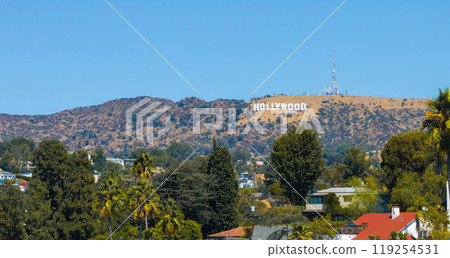 The iconic Hollywood Sign stands on Los Angeles hills, framed by clear skies. Lush trees and rooftops contrast with the rugged terrain in the foreground. The iconic Hollywood Sign stands on Los Angeles hills, framed by clear skies. Lush trees and rooftops contrast with the rugged terrain in the foreground. 119254531