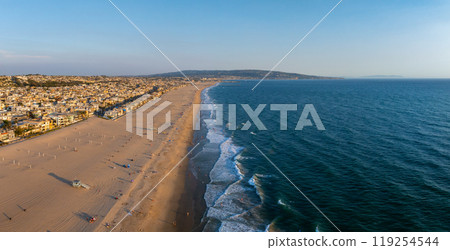 Aerial perspective of Manhattan Beach, California, featuring sandy shores, gentle waves, and residential buildings under a clear, sunlit sky. 119254544