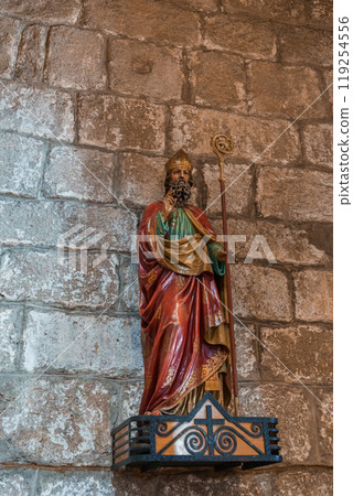 A detailed statue of a saint in vibrant robes with a staff, set against textured stone blocks, highlighting the historic charm of a Sardinian church. 119254556