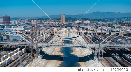 Aerial perspective of the Los Angeles River's concrete channel, featuring a large arched bridge, train tracks, urban buildings, and San Gabriel Mountains. Aerial perspective of the Los Angeles River's concrete channel, featuring a large arched bridge, train tracks, urban buildings, and San Gabriel Mountains. 119254630