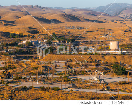 A vast desert landscape in California features numerous oil rigs, storage tanks, and power lines. Rolling hills and sparse vegetation are visible in the background. 119254631
