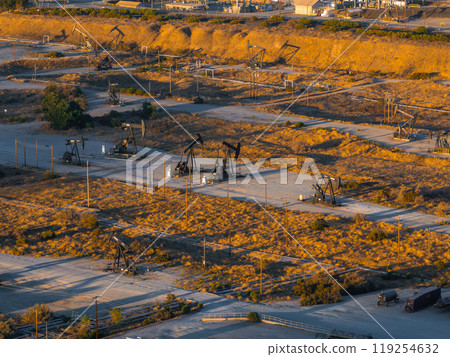 An aerial view captures numerous oil rigs in a California desert. The arid landscape features scrub vegetation and dirt roads, with long shadows cast by the setting sun. 119254632
