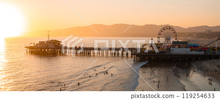 The Santa Monica Pier extends into the Pacific Ocean at sunset, featuring the illuminated Ferris wheel. People enjoy the beach with distant mountains visible. 119254633