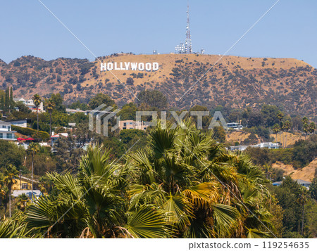 The iconic Hollywood Sign stands on Los Angeles hills, surrounded by rugged terrain and sparse vegetation. Palm trees and homes are visible in the foreground. 119254635