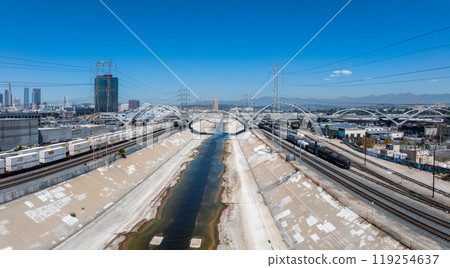 The Los Angeles River flows through a concrete channel, framed by the Sixth Street Viaduct. Downtown Los Angeles skyline and freight trains are visible. 119254637
