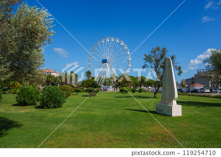 A lively park in Cagliari, Sardinia, featuring a large Ferris wheel, lush greenery, and a white stone monument under a clear blue sky. A lively park in Cagliari, Sardinia, featuring a large Ferris wheel, lush greenery, and a white stone monument under a clear blue sky. 119254710