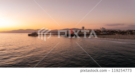 The Santa Monica Pier extends into the Pacific Ocean, featuring an illuminated Ferris wheel. The sunset casts warm hues over the ocean and cityscape. 119254773
