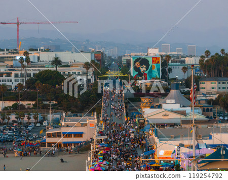 Santa Monica Pier in Los Angeles is crowded with people and vibrant umbrellas. A mural of a person with an afro adorns a building, with palm trees and a crane visible. Santa Monica Pier in Los Angeles is crowded with people and vibrant umbrellas. A mural of a person with an afro adorns a building, with palm trees and a crane visible. 119254792