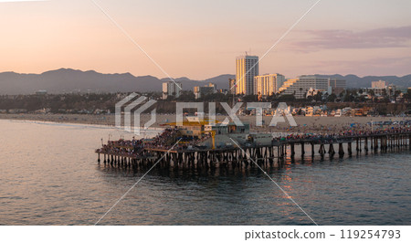 Santa Monica Pier extends into the Pacific Ocean, featuring a Ferris wheel and attractions. The cityscape and mountains are visible under a warm sunset glow. 119254793