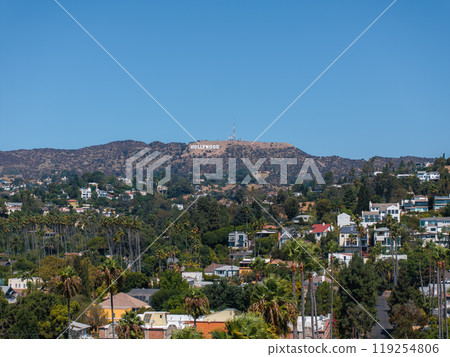 The iconic Hollywood Sign sits on the hills above Los Angeles, surrounded by lush greenery and residential buildings under a clear blue sky. 119254806