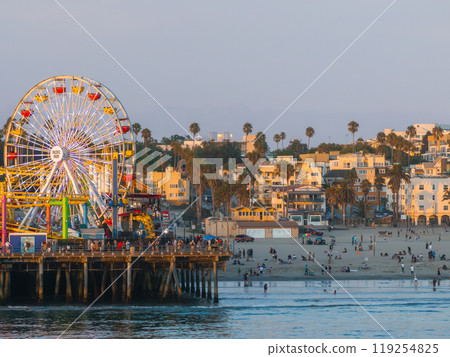 Santa Monica Pier features a vibrant Ferris wheel and amusement park. The beach is lively with people, palm trees, and coastal buildings in warm light. 119254825