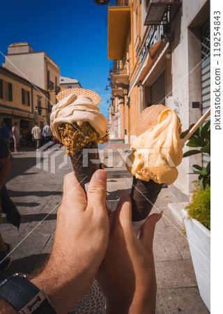 Two hands hold gelato cones with unique toppings against a vibrant street scene in Sardinia, Italy, featuring pastel buildings and a bright blue sky. 119254843
