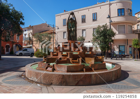 A tiered stone fountain with a bronze statue in a Sardinian square, surrounded by Mediterranean buildings and trees under a clear blue sky. A tiered stone fountain with a bronze statue in a Sardinian square, surrounded by Mediterranean buildings and trees under a clear blue sky. 119254858