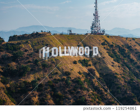 The Hollywood Sign is displayed on rugged hills with a communications tower. Distant mountains and a clear blue sky form the scenic backdrop. 119254861