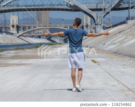 A person stands with arms outstretched on the concrete banks of the Los Angeles River, with multiple arched bridges and urban buildings in the background. 119254862
