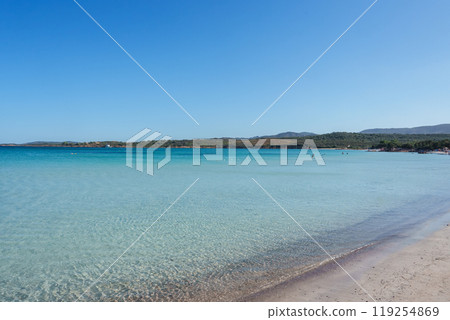 Experience tranquility on a pristine Sardinian beach with crystal clear waters, sandy shores, and a picturesque backdrop of hills under a bright blue sky. Experience tranquility on a pristine Sardinian beach with crystal clear waters, sandy shores, and a picturesque backdrop of hills under a bright blue sky. 119254869