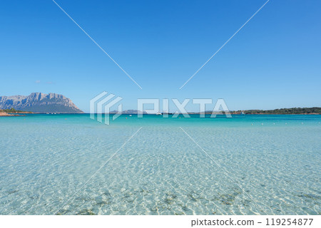 Experience the tranquil beauty of Sardinia with crystal clear waters, a sailboat, and a rugged mountain backdrop under a cloudless sky. 119254877