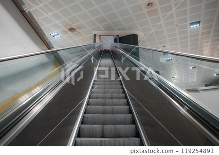 Upward view of a sleek escalator with glass railings and grid lighting, showcasing contemporary architecture in a Sardinian shopping mall or airport. Upward view of a sleek escalator with glass railings and grid lighting, showcasing contemporary architecture in a Sardinian shopping mall or airport. 119254891