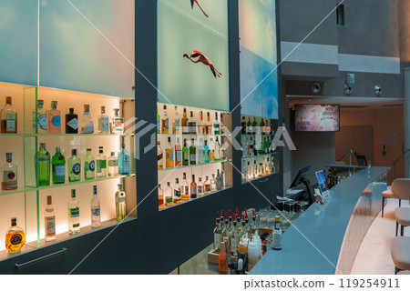 Discover a sophisticated bar in a luxury Sardinian hotel, featuring a curved counter, illuminated liquor shelves, and unique bird wall decorations. 119254911