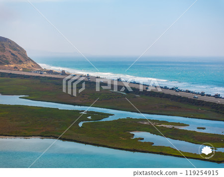 Aerial view of San Diego's coastal area featuring a beach, Pacific Ocean, lush wetlands, and a road with vehicles. A hill is visible in the background. 119254915