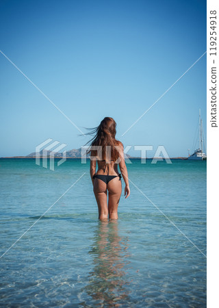 A serene scene of a woman in shallow waters at a Sardinian beach, with a sailboat and rocky hills in the background, under a bright blue sky. A serene scene of a woman in shallow waters at a Sardinian beach, with a sailboat and rocky hills in the background, under a bright blue sky. 119254918