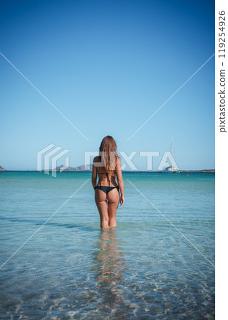 A serene scene in Sardinia featuring a woman standing in clear waters, with a white sailboat anchored in the distance under a clear blue sky. A serene scene in Sardinia featuring a woman standing in clear waters, with a white sailboat anchored in the distance under a clear blue sky. 119254926