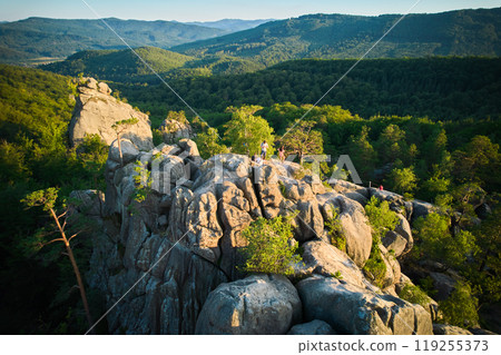 Aerial view of prominent rock formation amidst dense forest. Rolling hills and distant mountain ranges create breathtaking backdrop. Dovbush Rocks, Carpathian mountains, Ukraine. 119255373