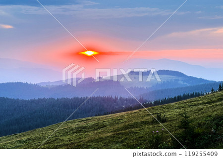 Sun sets behind range of silhouetted mountains, casting warm, golden glow across sky. Soft clouds, tinged with hues of pink and orange, drift lazily, creating serene and tranquil evening scene. 119255409