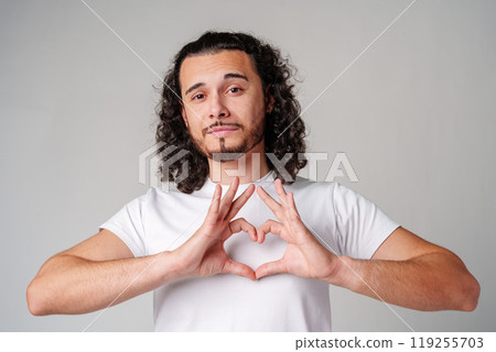 Young man with curly hair forms a heart shape with his hands while standing against a light background Young man with curly hair forms a heart shape with his hands while standing against a light background 119255703