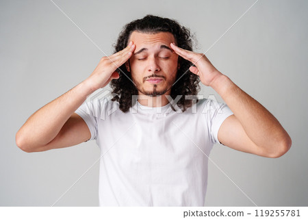 A young man with curly hair rubs his forehead in a casual indoor setting while expressing frustration A young man with curly hair rubs his forehead in a casual indoor setting while expressing frustration 119255781