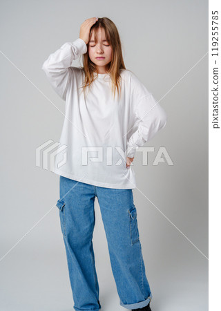 Young girl in a white sweater holding her head in discomfort against a minimalist background Young girl in a white sweater holding her head in discomfort against a minimalist background 119255785