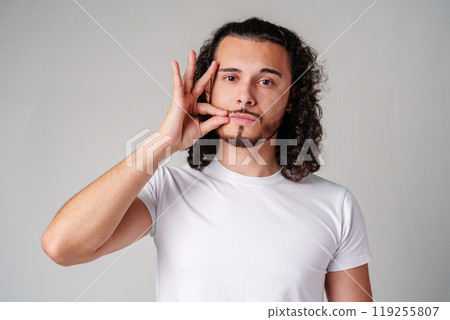 A young man with curly hair demonstrates a hand gesture in a plain white shirt against a neutral background A young man with curly hair demonstrates a hand gesture in a plain white shirt against a neutral background 119255807