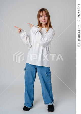 Young girl happily pointing against a neutral background while wearing a cozy white sweatshirt 119255825