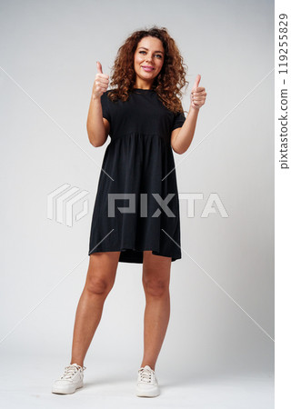 Cheerful young woman with curly hair giving a thumbs up while smiling against a neutral background 119255829