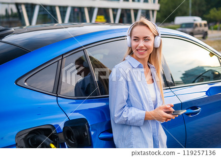Electric car charging at a station with a woman listening to music via headphones using her phone. 119257136