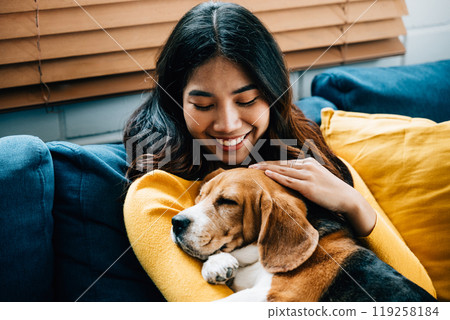 In their living room, an Asian woman and her cute Beagle puppy share a heartwarming nap on the sofa, epitomizing the concept of trust, togetherness, and happiness. It's a portrait of love at home. In their living room, an Asian woman and her cute Beagle puppy share a heartwarming nap on the sofa, epitomizing the concept of trust, togetherness, and happiness. It's a portrait of love at home. 119258184