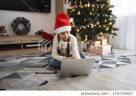 Young girl in Santa hat using laptop by Christmas tree Young girl in Santa hat using laptop by Christmas tree 119258667