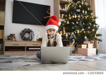Young girl using laptop by Christmas tree in festive living room Young girl using laptop by Christmas tree in festive living room 119258668