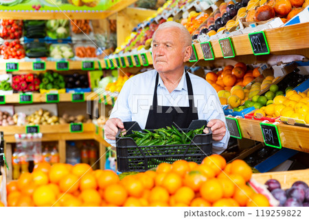 Elderly supermarket employee rearranges box of green chili peppers 119259822