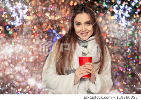 Girl in fur coat stands with glass of coffee in hands on shiny flashing festive sparkling background 119260055