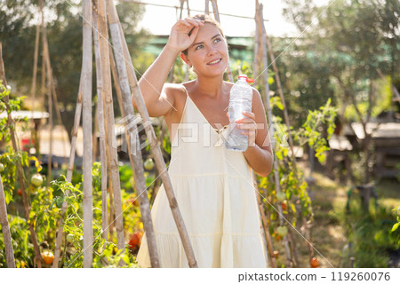 Girl stands near garden bed and drinks water from bottle 119260076