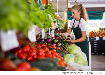 Young girl who works part-time as a trainee saleswoman puts on the showcase eggplants Young girl who works part-time as a trainee saleswoman puts on the showcase eggplants 119260135