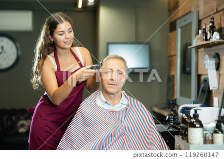 Young female barber making haircut to elderly client in barbershop Young female barber making haircut to elderly client in barbershop 119260147