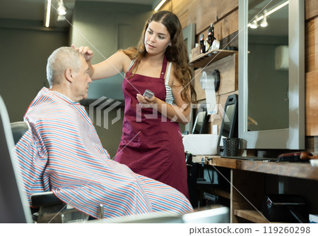 Young female barber discussing haircut with elderly client in barbershop Young female barber discussing haircut with elderly client in barbershop 119260298