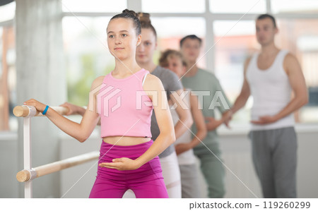 Teenagers doing ballet at group training session in the studio performs demi plie near the ballet barre, standing in ballet stance Teenagers doing ballet at group training session in the studio performs demi plie near the ballet barre, standing in ballet stance 119260299