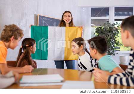 Inspired young female teacher conducting lesson for preteens, showing state flag of Ireland in audience of middle school Inspired young female teacher conducting lesson for preteens, showing state flag of Ireland in audience of middle school 119260533