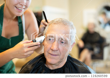 Elderly man getting haircutting by female barber using clipper in barbershop 119260599
